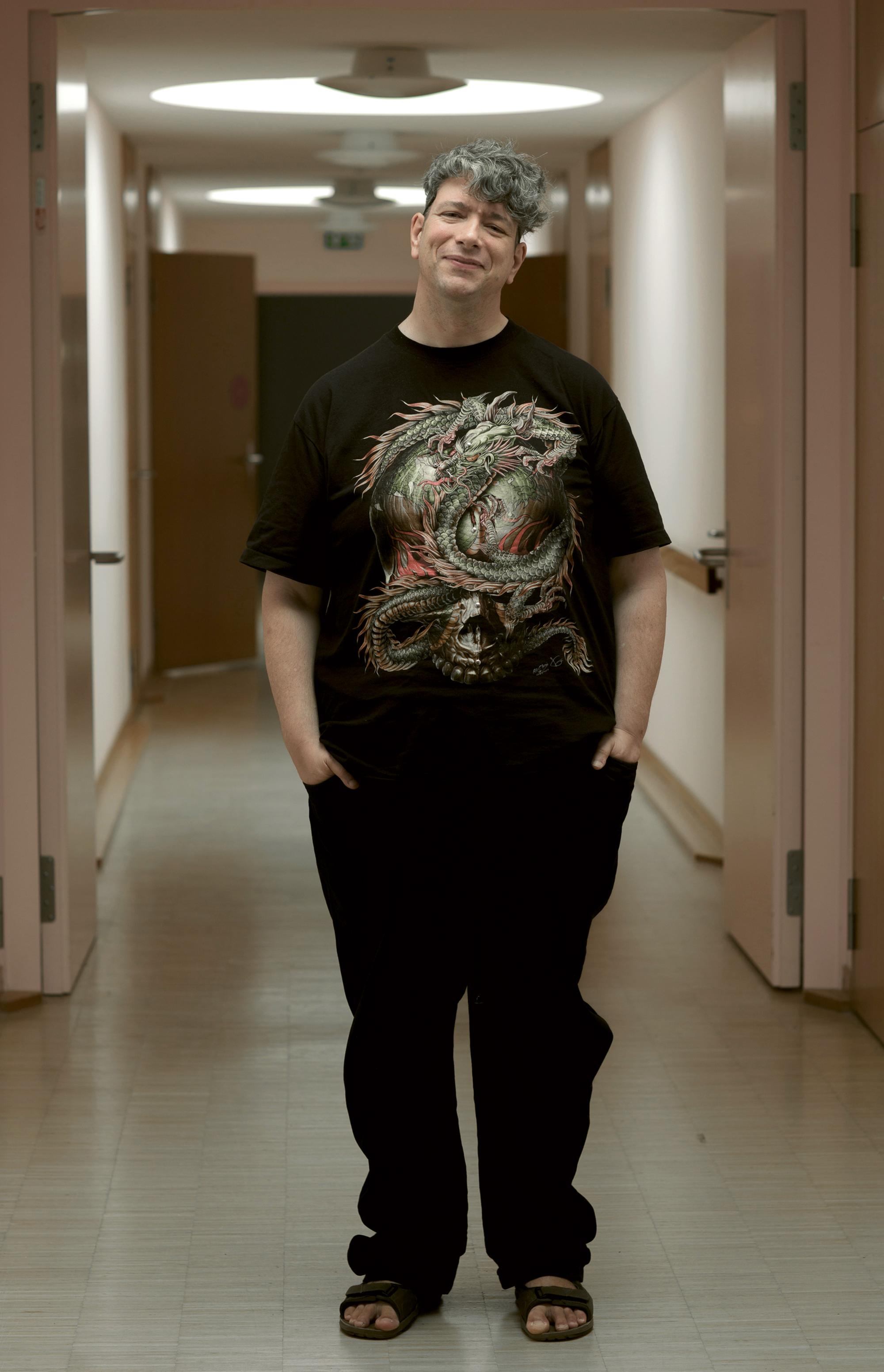 Ein Ganzkörperfoto von Remo Beuggert, einem weißen Mann mit Behinderung. Remo steht in einem hell erleuchteten Gang. Hinter ihm sind verschiedene Türen, teils offen, teils geschlossen. Remos Kopf ist leicht zur Seite geneigt und er schmunzelt. Seine Hände hat er in seine Hosentaschen gesteckt. Er hat graues gewelltes kurzes Haar und trägt ein schwarzes T-Shirt mit einem großen bunten Drachenaufdruck sowie schwarze Hosen und Sandalen. Foto: Ona Pinkus