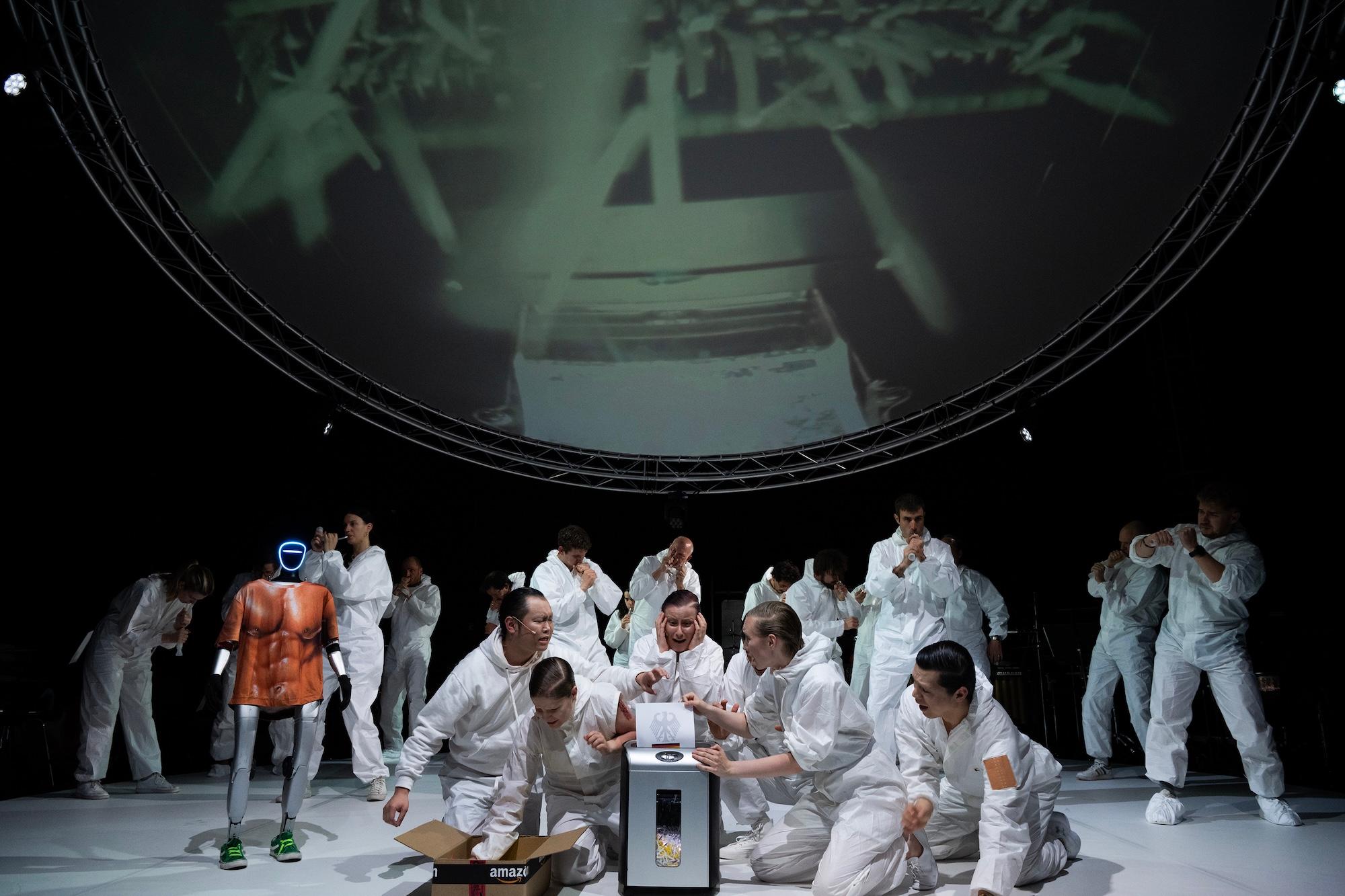 Zum Doppeljubiläum von Hans Werner Henze und der Münchner Biennale lässt Piyawat Louilarpprasert in „R3SIST4NC3 B0D1EZ“ Henzes politisch aufgeladene „Voices“-Lieder auf forensische Laborszenen, Roboterin Karla, Fast-Fashion-Fließbänder, TikTok-Feeds und „Make Klassenkampf great again“-Raves prallen.